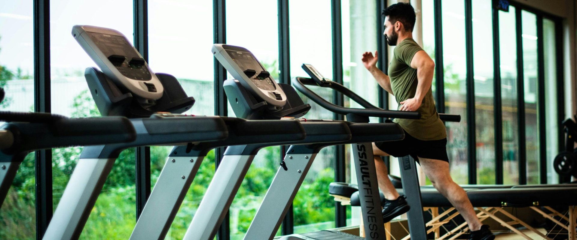 Young man workouts on treadmill in modern gym with large windows and natural light.